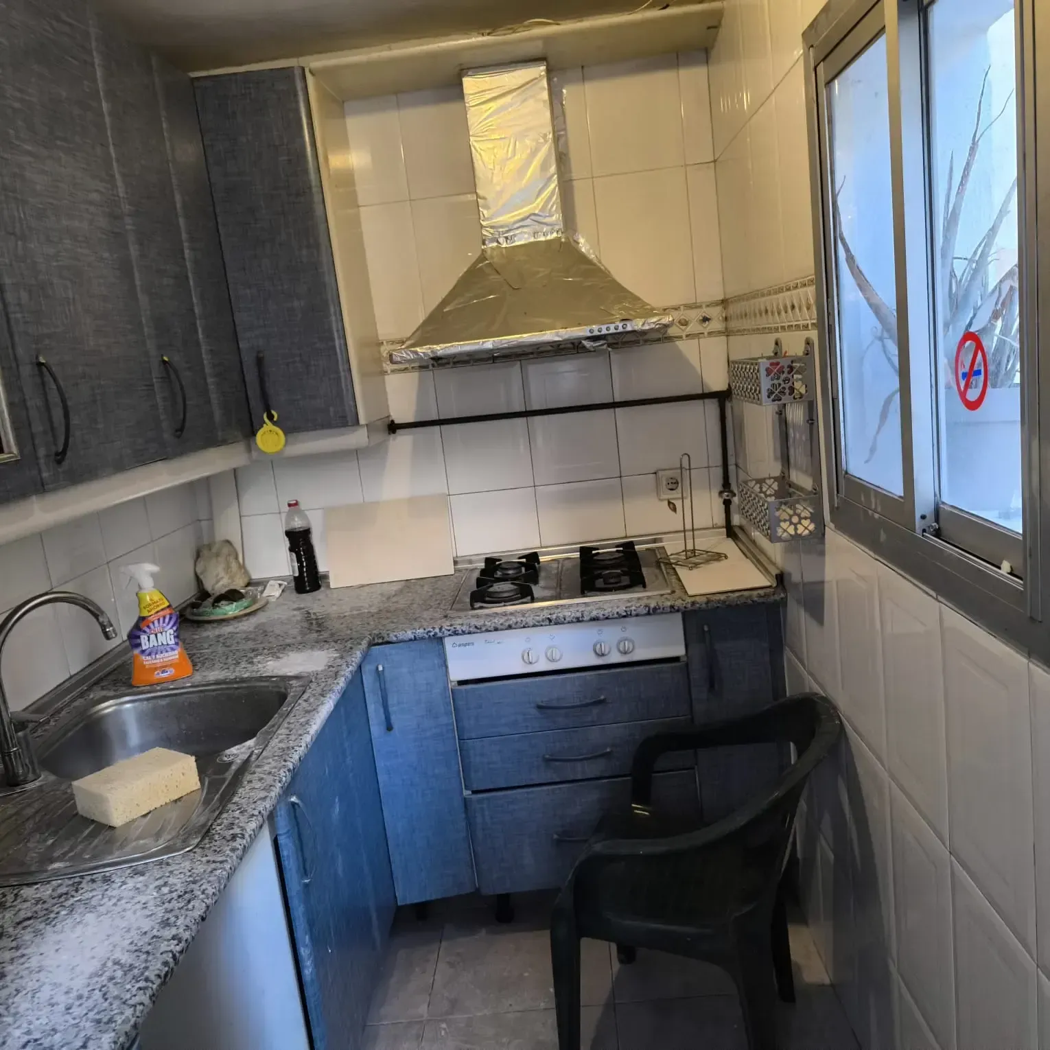 Cocina pequeña y estrecha con armarios grises, paredes alicatadas, fregadero, fogones y una ventana a la derecha.