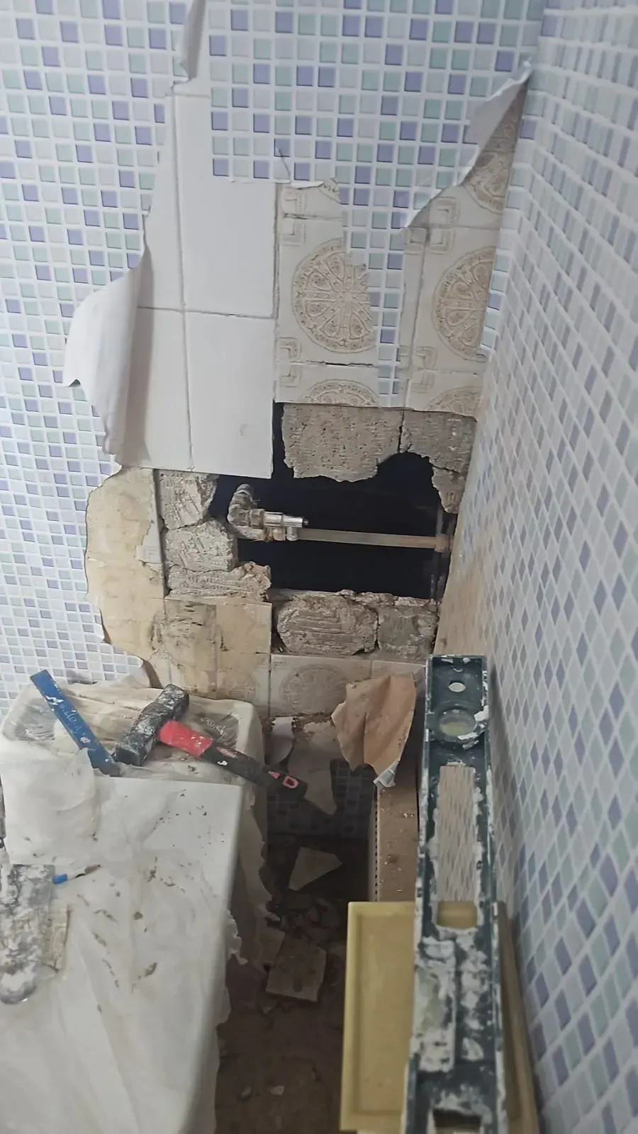 Pared del baño dañada con un gran agujero que deja al descubierto las tuberías y escombros en el suelo.