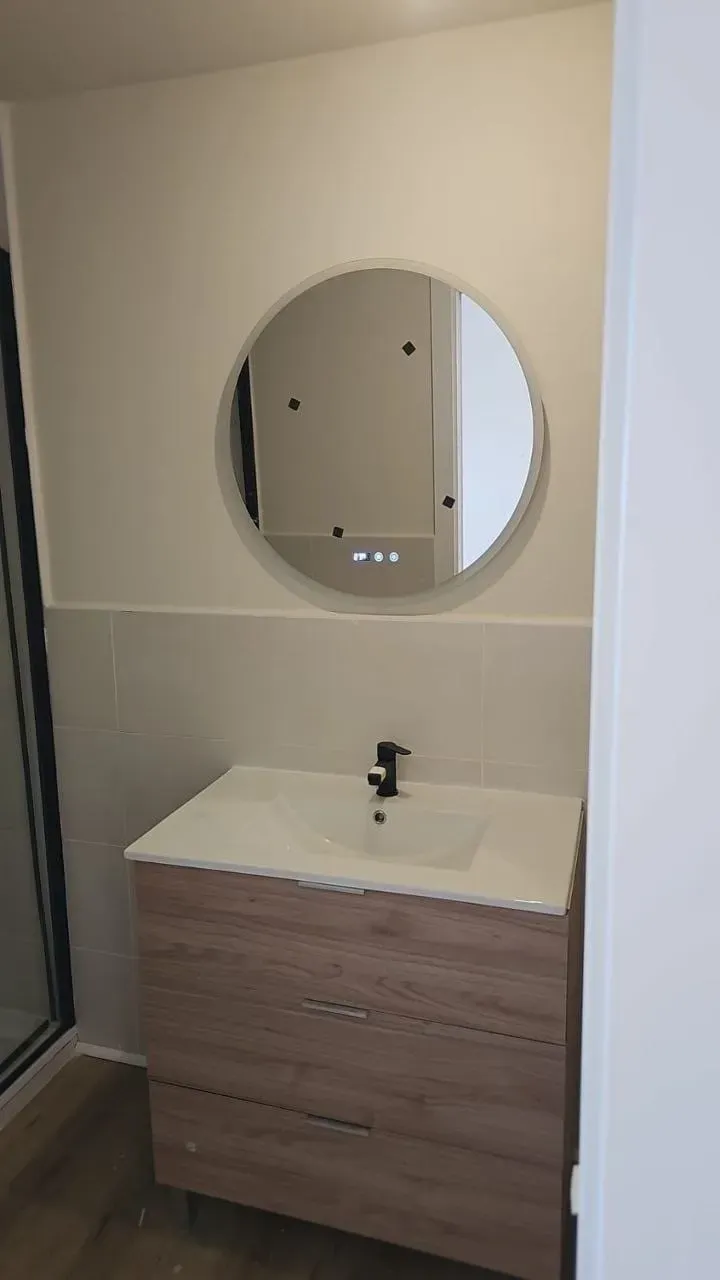 Pequeño tocador de baño con espejo redondo, lavabo blanco y mueble de madera sobre paredes de color beige.