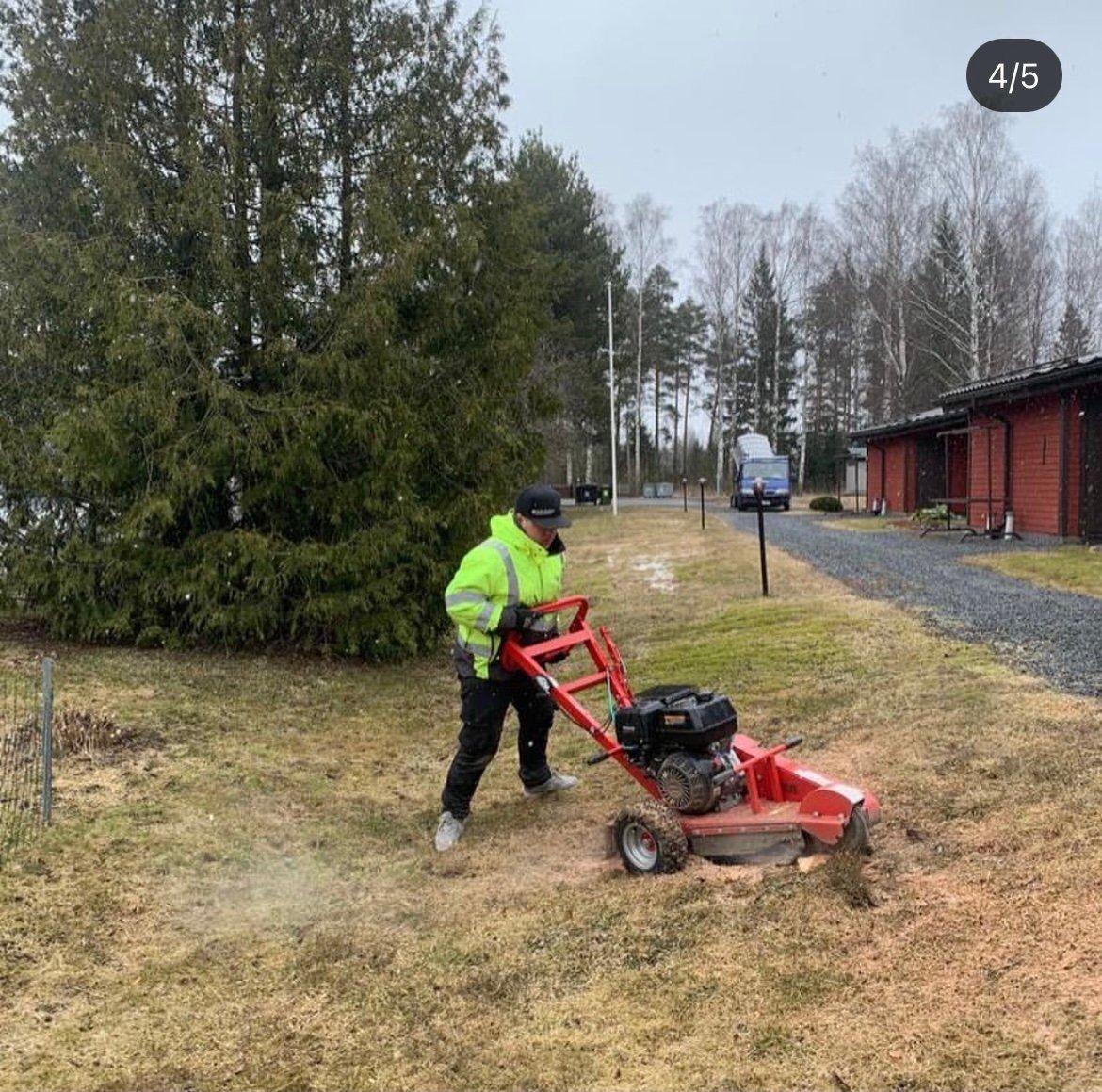 Neonväriseen takkiin pukeutunut henkilö leikkaa kuivaa ruohoa punaisen mökin ja sorapolun vieressä.