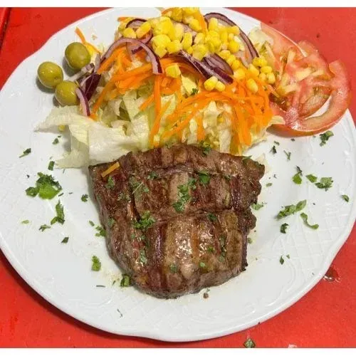 Filete a la parrilla con ensalada, aceitunas y rodajas de tomate en un plato blanco.