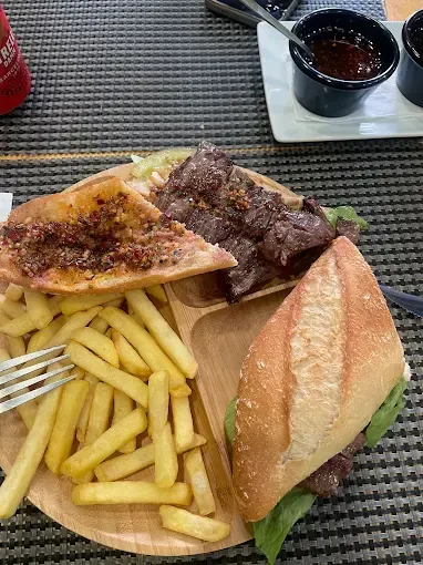 Plato de patatas fritas, un sándwich tostado y carne a la parrilla en una tabla de madera con un vasito para salsa.
