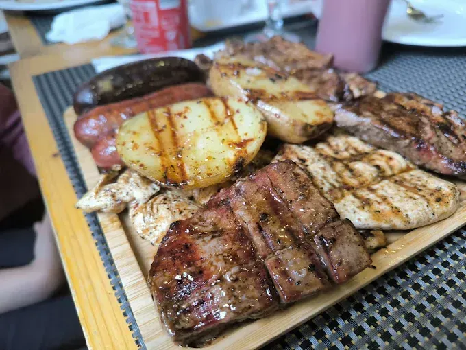 Carne y verduras a la parrilla en una bandeja, incluyendo bistec, salchicha y patatas, con bebidas al fondo.
