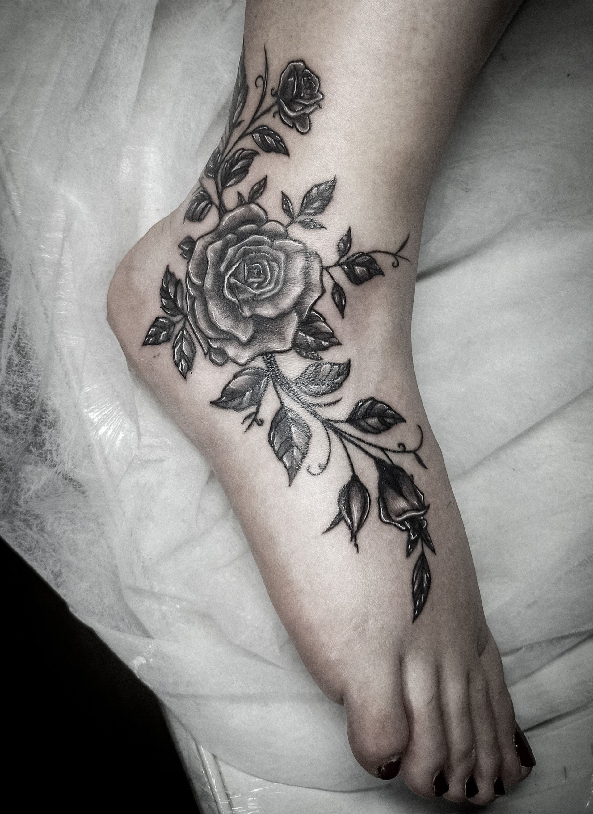 Tatuaje de rosa negra en el tobillo, enredaderas que se extienden hacia arriba y hacia abajo del pie, sombreado detallado.