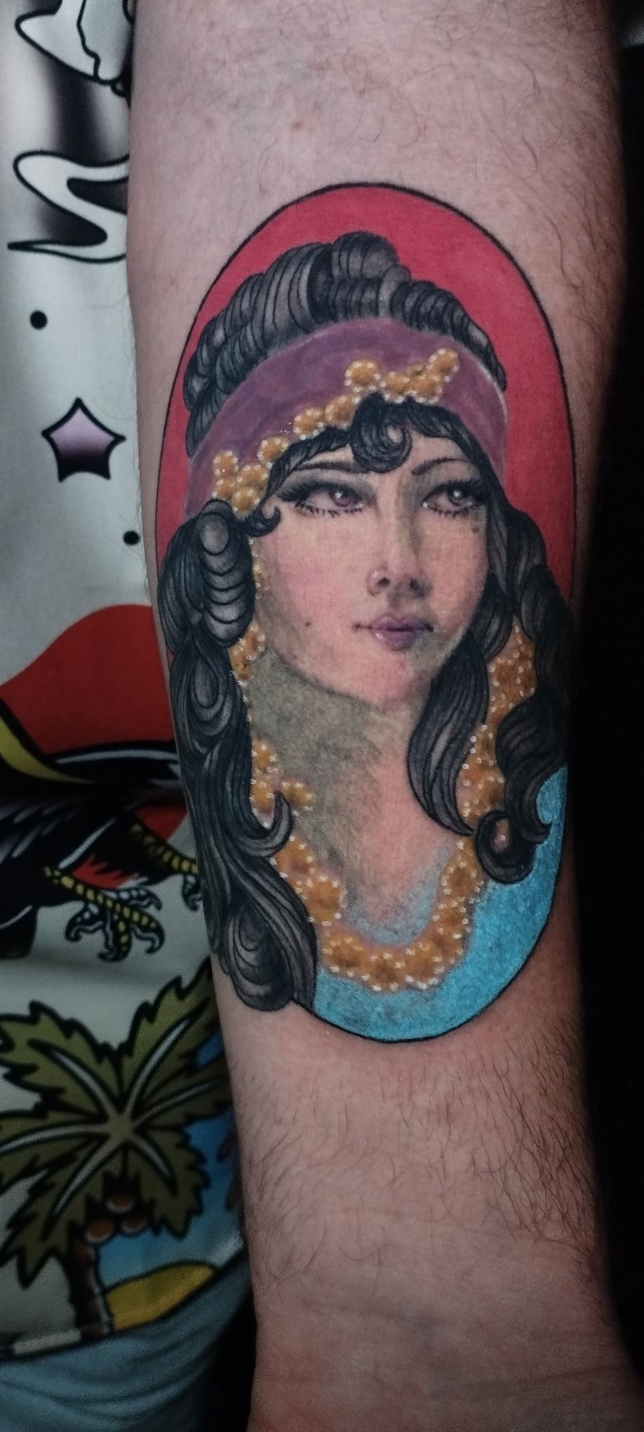 Tatuaje de una mujer con pañuelo morado, collar dorado y vestido turquesa. Fondo rojo, en el brazo.