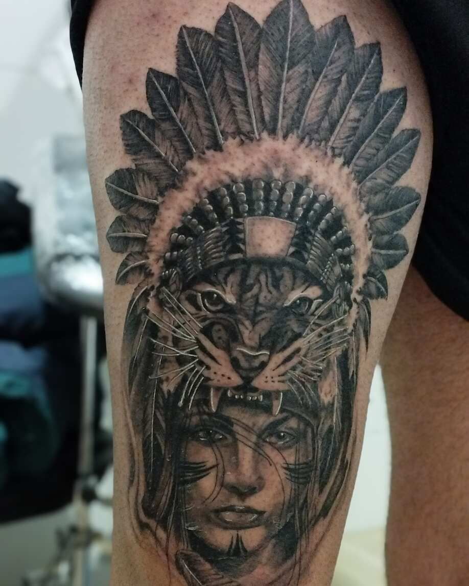 Tatuaje del rostro de una mujer debajo de un tocado de tigre y un tocado de plumas, en negro y gris en una pierna.
