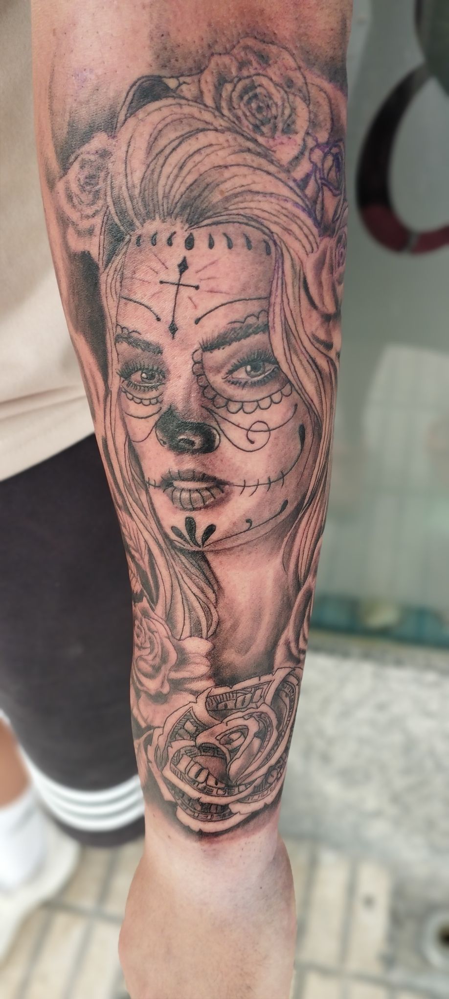 Tatuaje de un rostro de mujer al estilo de una calavera de azúcar en un brazo, rodeado de rosas.
