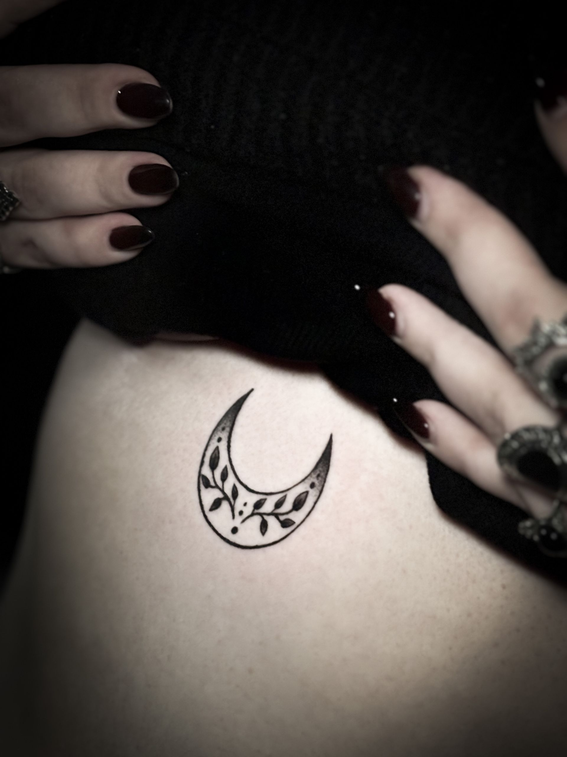 Tatuaje de luna creciente con hojas en el costado de una persona, manos en la parte superior; uñas oscuras y anillos.