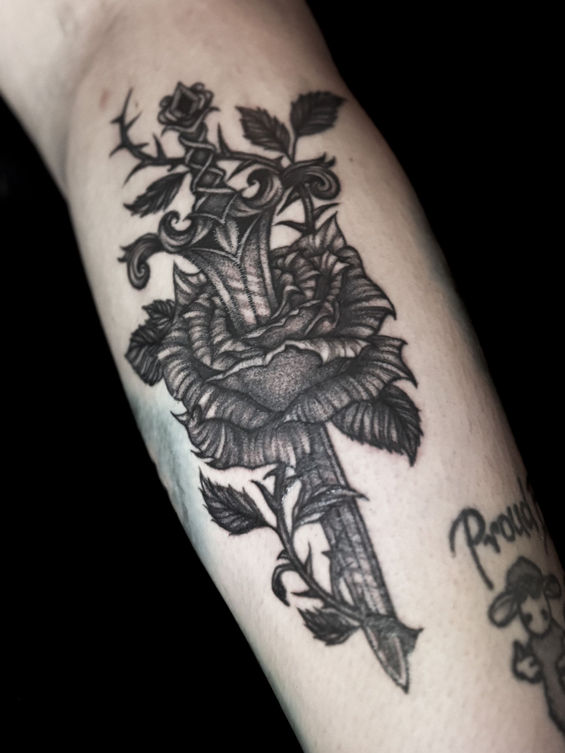 Tatuaje de una daga perforando una gran rosa, rodeada de espinas y hojas, en un antebrazo.