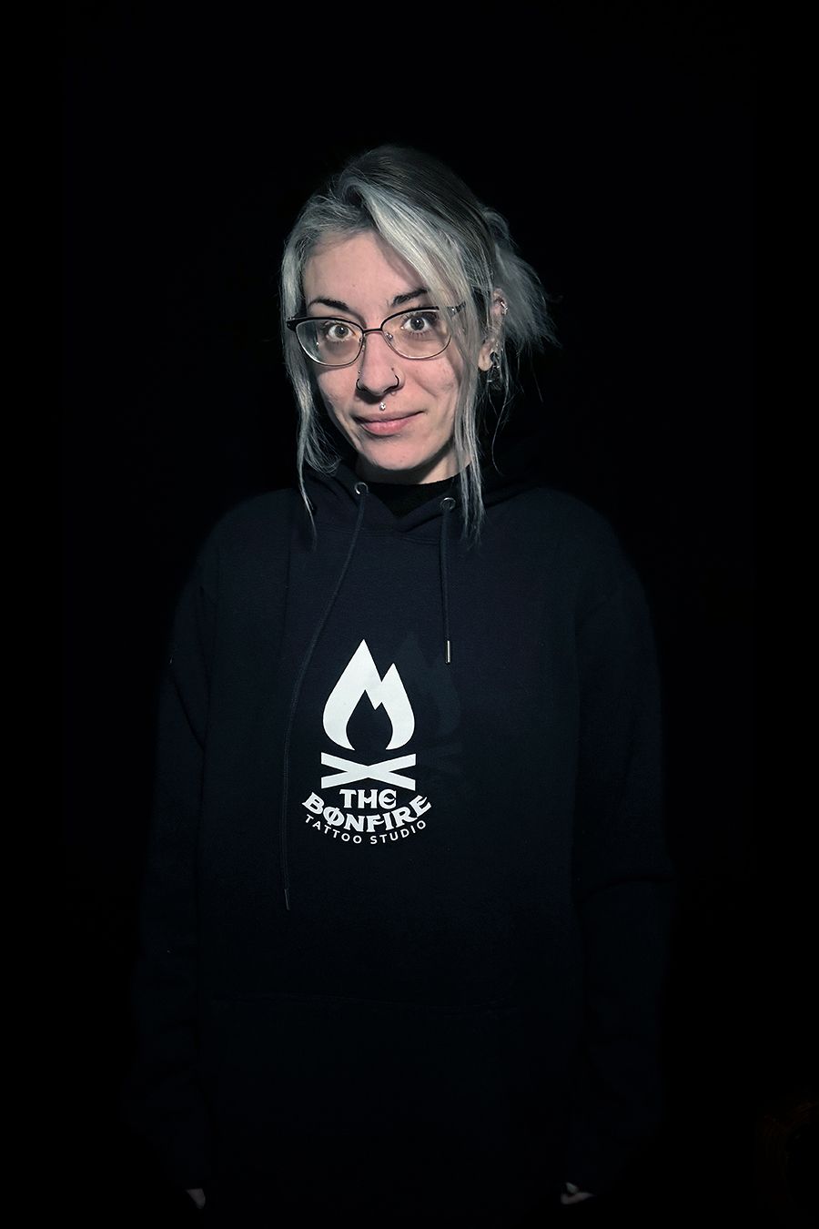 Mujer con gafas y cabello gris, vestida con una sudadera con capucha negra con el logo de una fogata, sobre un fondo negro.
