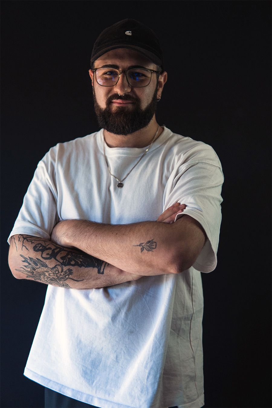 Hombre con gafas, barba y gorra de béisbol, brazos cruzados, tatuajes visibles sobre fondo negro.