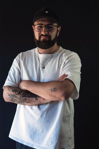 Un hombre con gafas, barba y gorra, con los brazos cruzados, está de pie frente a un fondo negro.