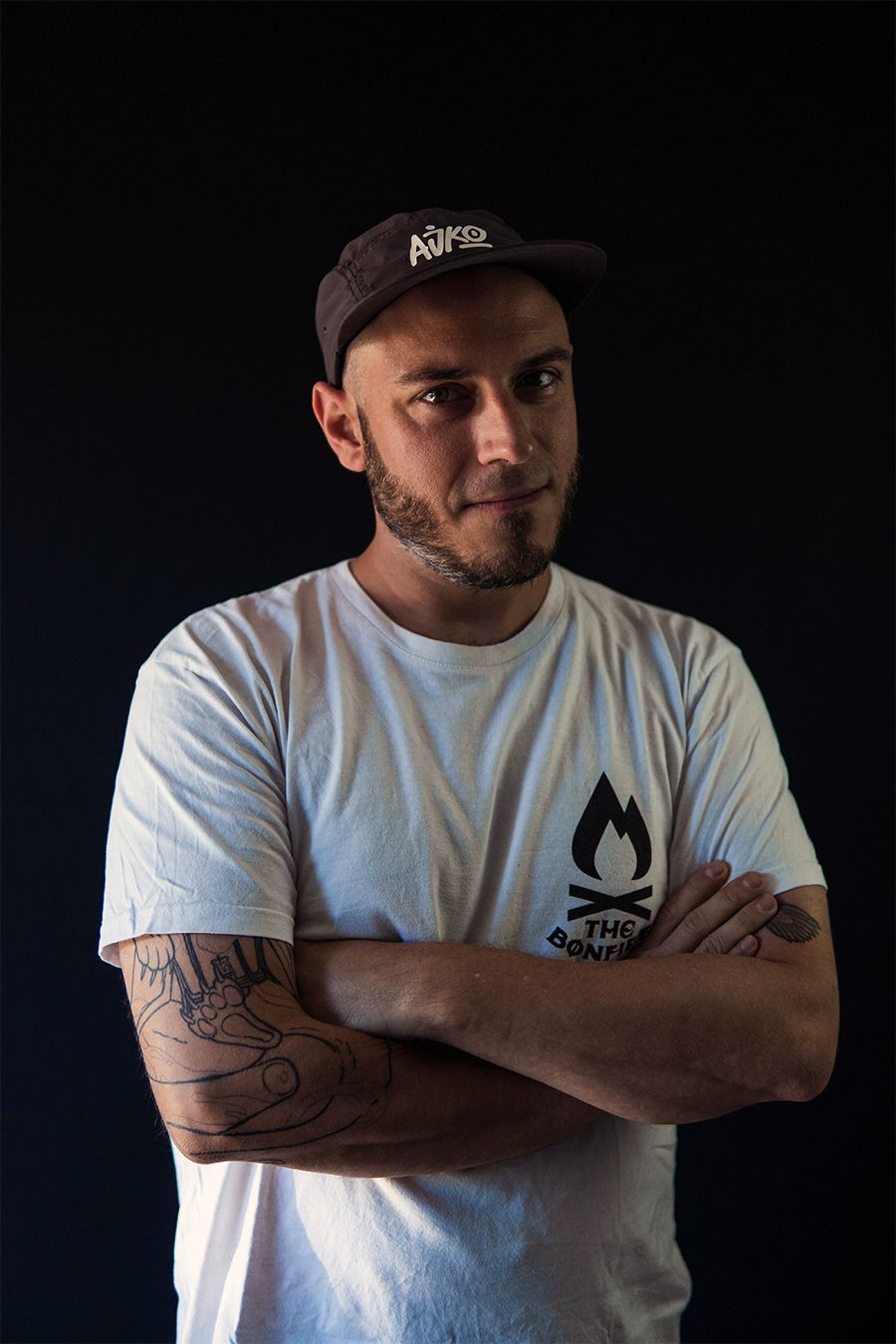 Hombre con los brazos cruzados, cabeza rapada, perilla, vestido con camiseta blanca, sombrero negro y tatuajes, delante de un fondo negro.