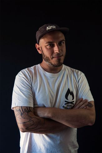 Hombre con los brazos cruzados, con gorra y camiseta con el logo de una fogata, tatuajes, sonriendo.
