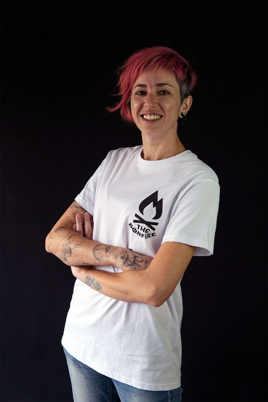 Mujer con cabello rosa, brazos cruzados, sonriente, vistiendo una camiseta blanca con el logo de una hoguera, jeans, parada frente a un fondo negro.