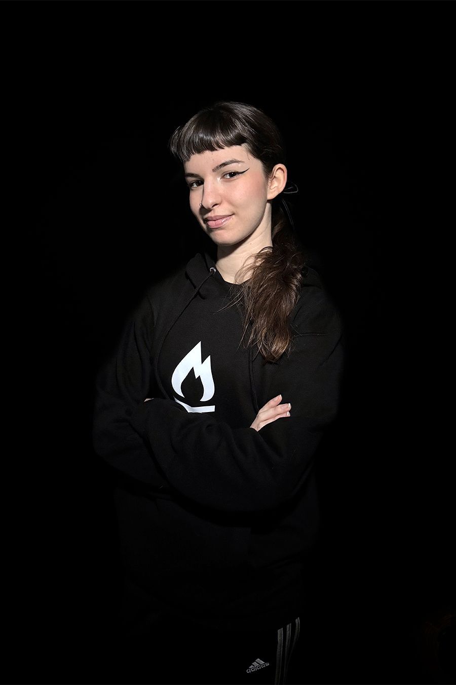 Mujer con los brazos cruzados, vestida con una sudadera negra con el logo de una llama blanca y fondo oscuro.