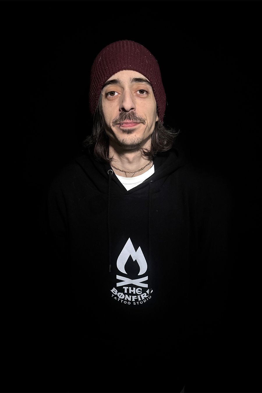 Hombre que viste un gorro y una sudadera con capucha negra con el logo de una hoguera, sonriendo sobre un fondo oscuro.