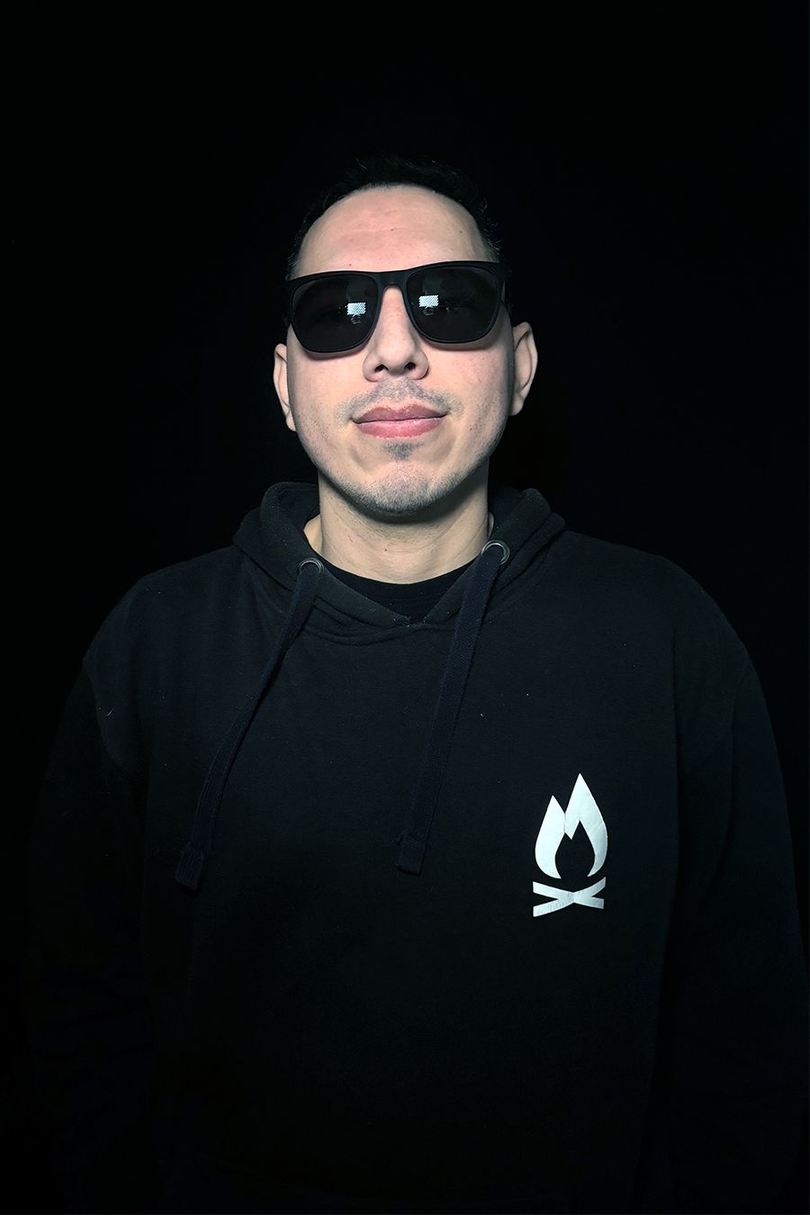 Hombre con gafas de sol y sudadera con capucha negra con el logotipo de una fogata blanca y fondo negro.