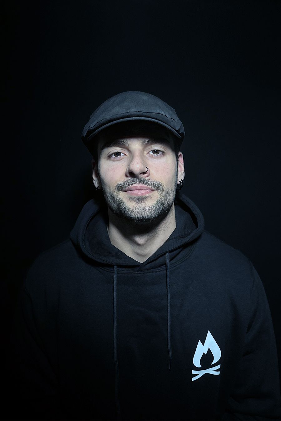 Hombre con sudadera negra y gorra de repartidor de periódicos, sonriendo a la cámara sobre un fondo oscuro. Se ve el logo blanco de una hoguera.