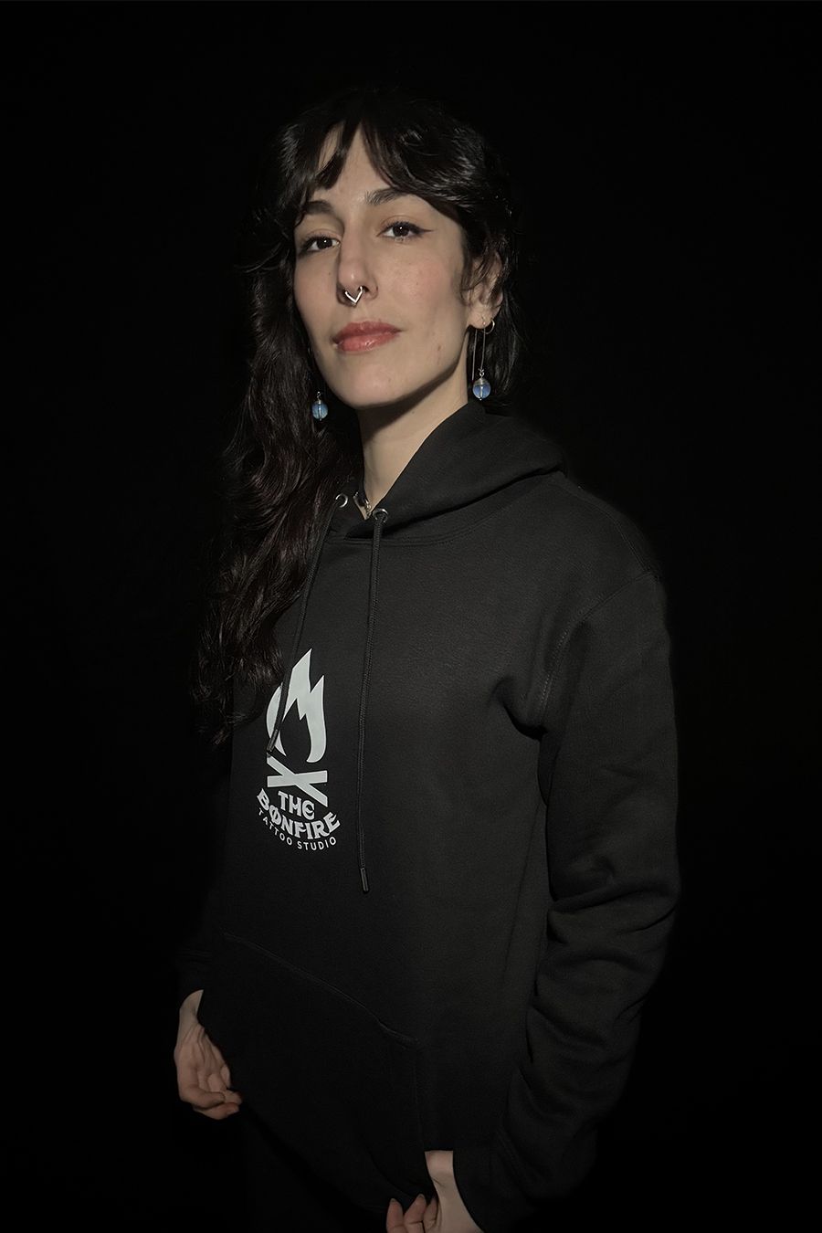 Mujer con sudadera con capucha negra y gráfico de fogata, de pie sobre un fondo oscuro.