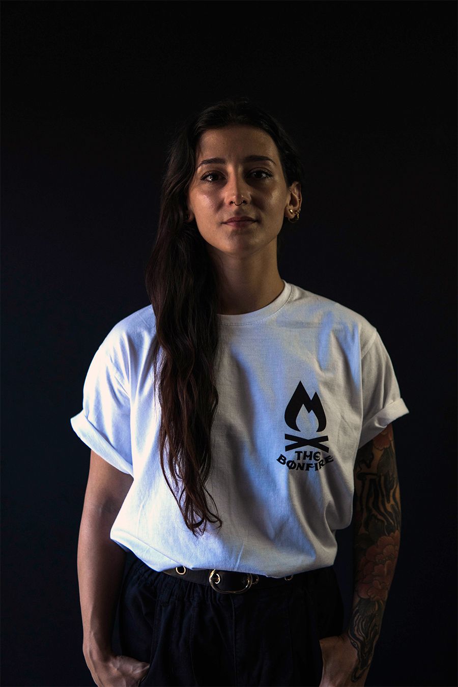 Mujer con cabello oscuro, vestida con una camiseta blanca con el logo de una llama, pantalones negros y tatuajes en el brazo, sobre un fondo negro.