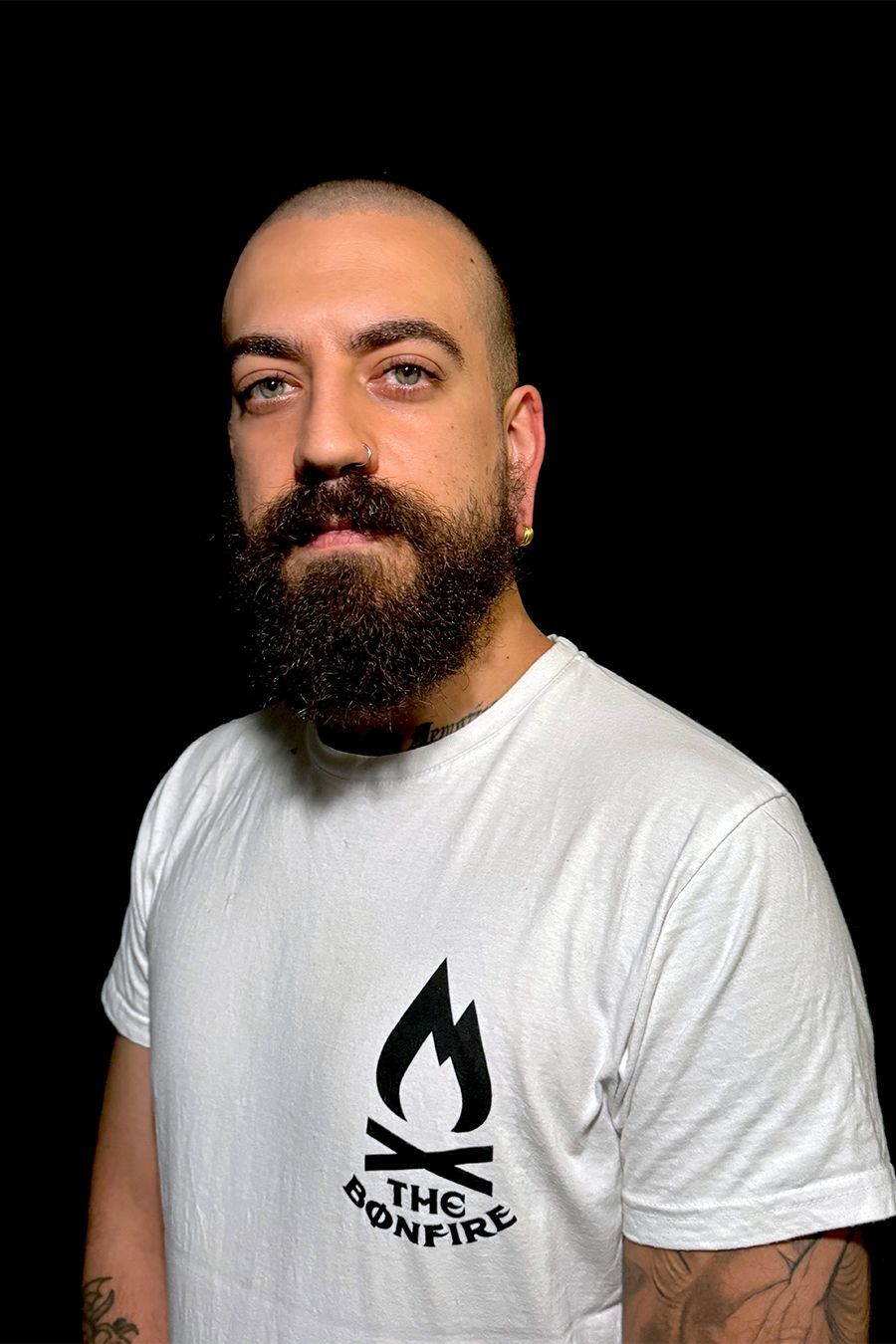 Hombre con barba y cabeza rapada, vestido con una camiseta blanca con el logo de una hoguera, mirando a la cámara.