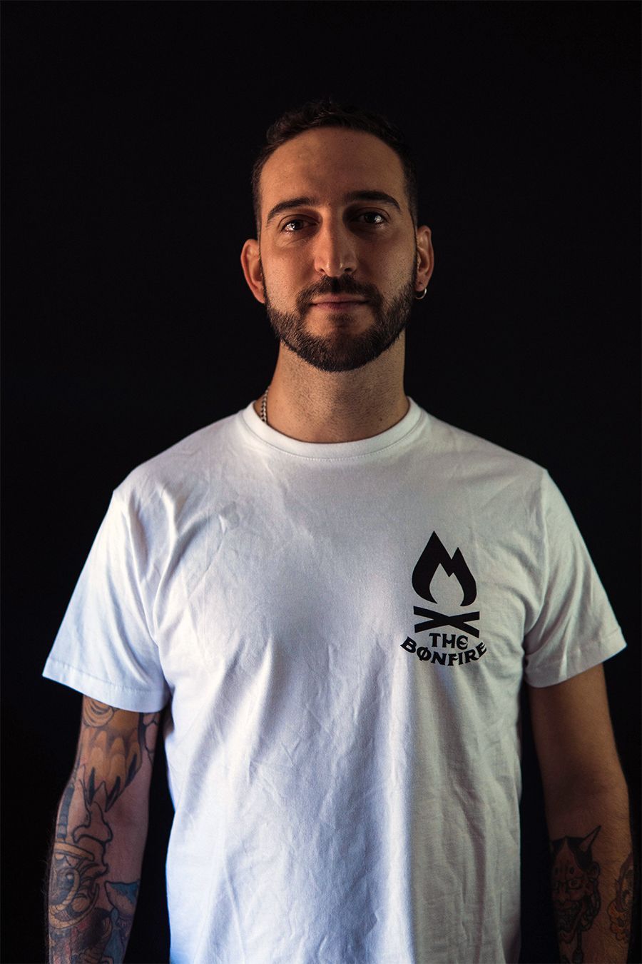 Hombre con barba, camiseta blanca con el logo de una hoguera y brazos tatuados sobre un fondo negro.