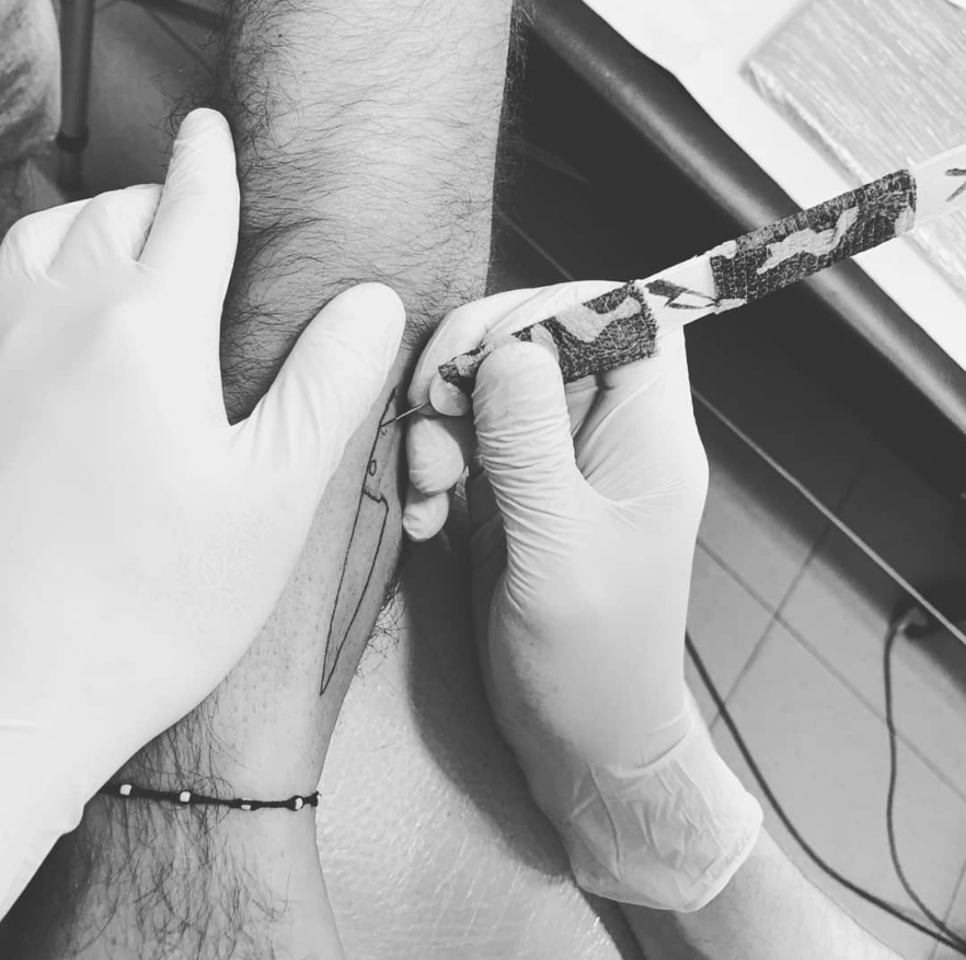 El antebrazo de una persona siendo tatuado con un diseño de cuchillo por alguien que usa guantes, en un lugar interior.