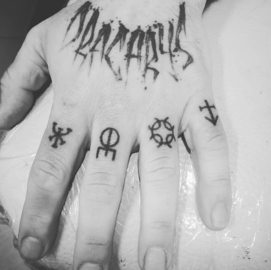 Mano con múltiples tatuajes, incluyendo símbolos en los dedos y una palabra en el dorso. Blanco y negro.