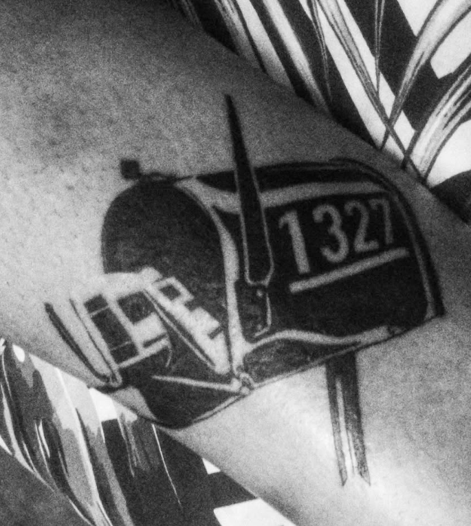 Tatuaje en blanco y negro de un buzón con el número 1327 en el brazo de una persona.