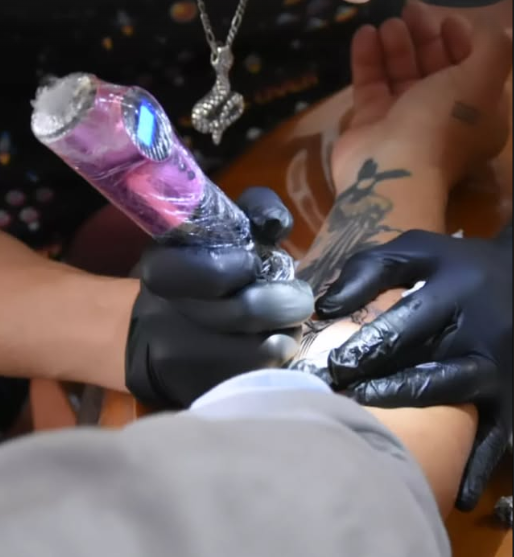 Un tatuador con guantes negros entinta un diseño en el antebrazo de una persona. La máquina de tatuar es iridiscente.