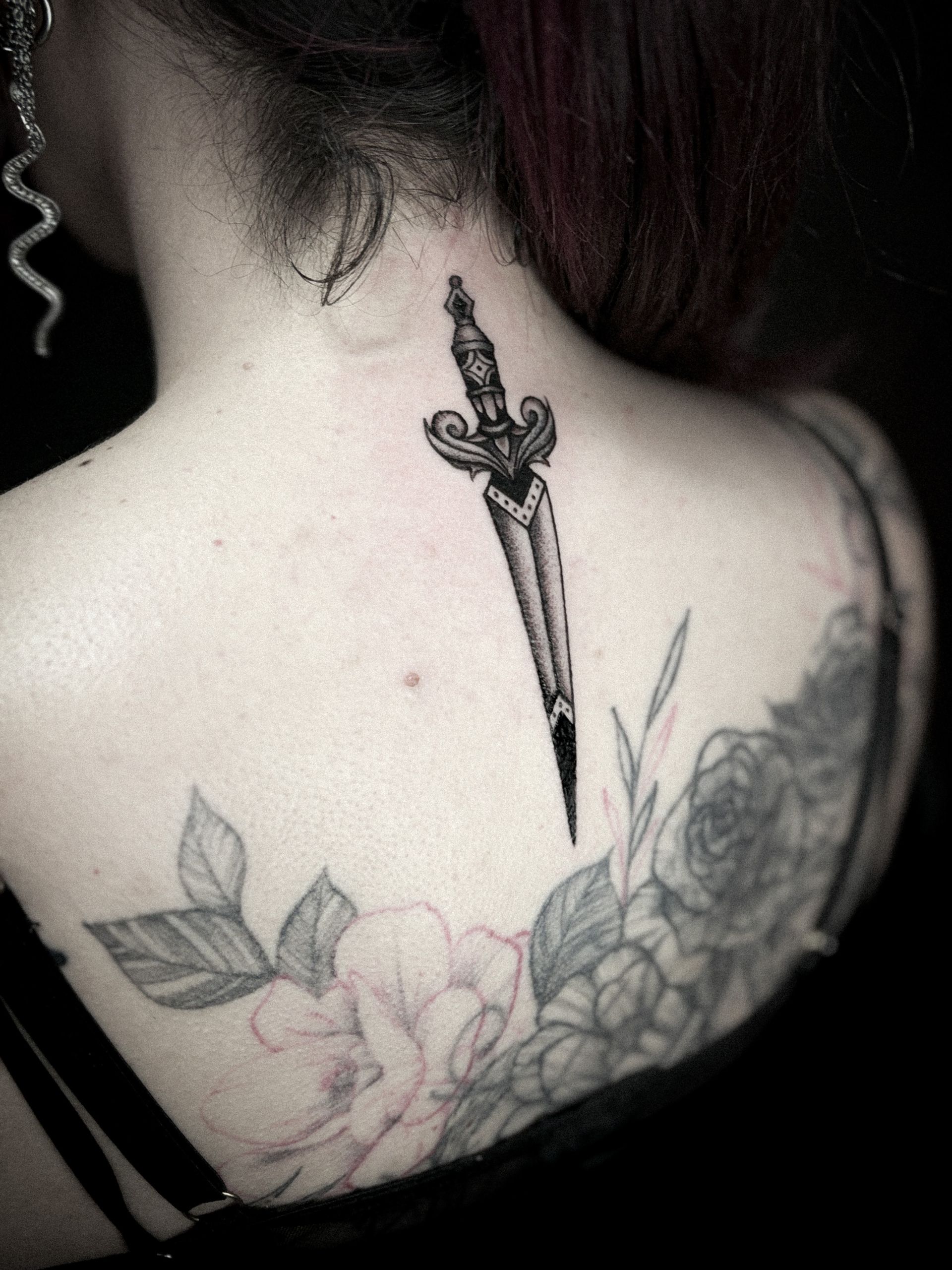 Espalda de mujer con un tatuaje de daga en el cuello y un tatuaje floral a lo largo del hombro.