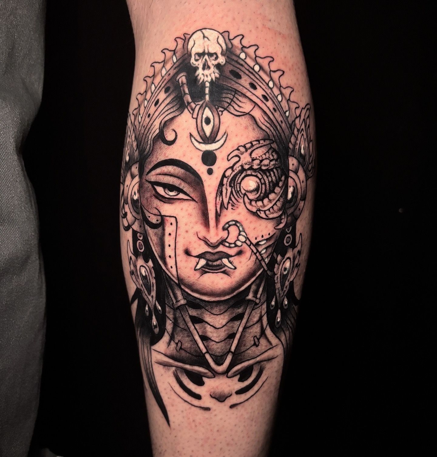 Tatuaje en blanco y negro del rostro de una deidad, un lado detallado y el otro tipo cyborg con una calavera.