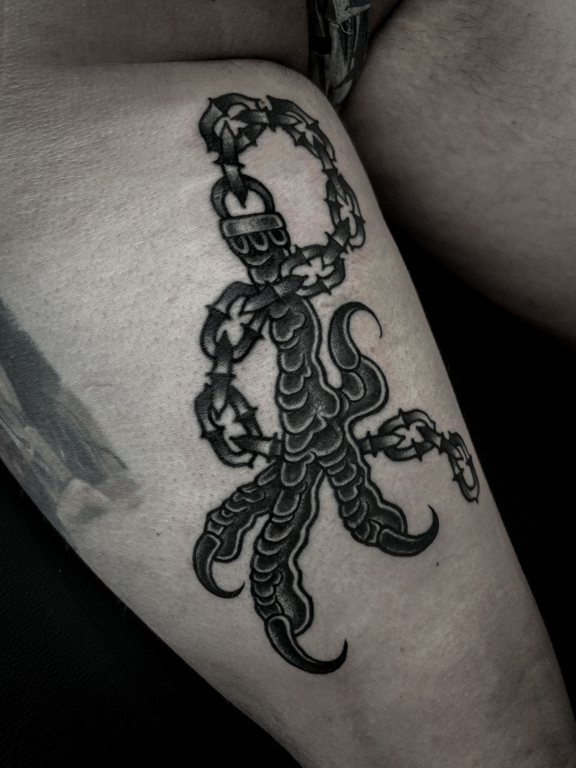 Tatuaje negro y gris de un pie con garras y una cadena con púas, en el muslo de una persona.