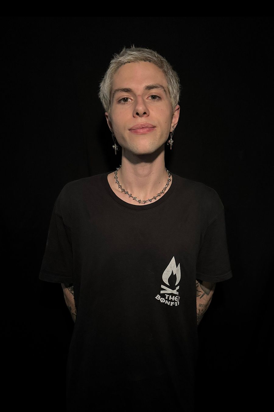 Hombre con barba, camiseta blanca con el logo de una hoguera y brazos tatuados sobre un fondo negro.
