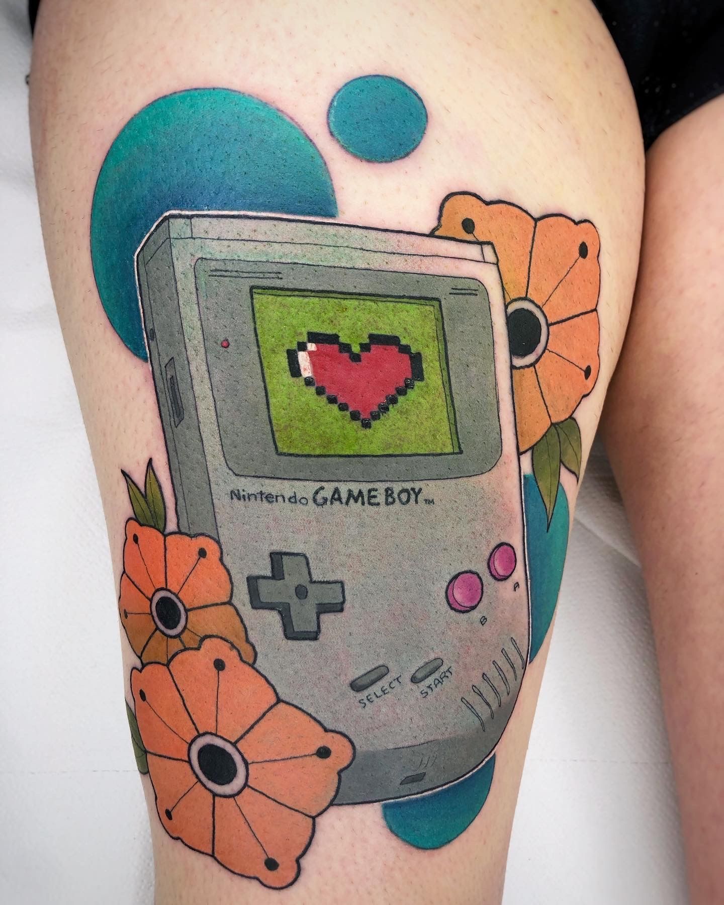 Tatuaje de un Game Boy gris con un corazón pixelado en la pantalla, flores naranjas y círculos verde azulado en la piel.
