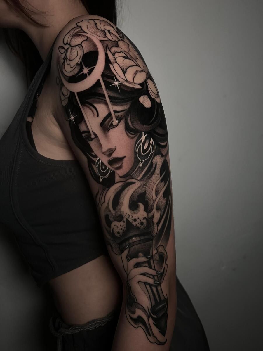 Tatuaje en el brazo de una mujer que presenta una figura femenina celestial sosteniendo una antorcha bajo una luna creciente, realizado en negro y gris.