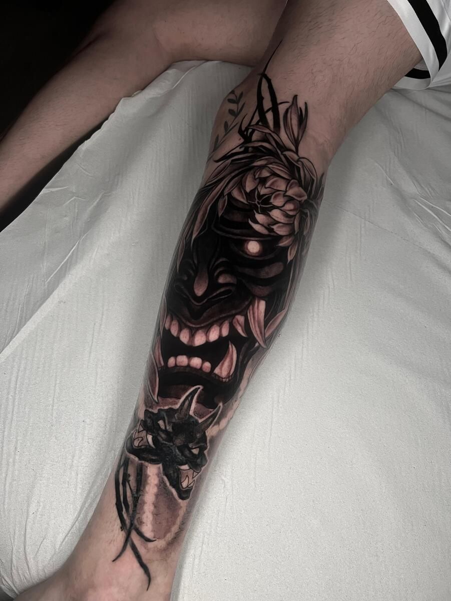 Tatuaje de máscara Hannya negra y gris en la pierna de una persona, con elementos florales y sombreado.