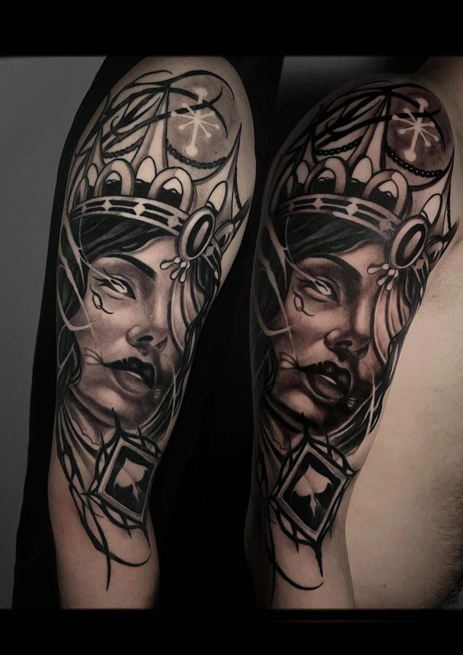 Tatuaje negro y gris del rostro de una reina en el brazo. Lleva una corona y un colgante.