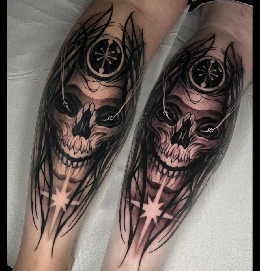 Dos tatuajes en el antebrazo: una calavera estilizada con un objeto parecido a una brújula encima y una estrella debajo, en tinta negra y gris.