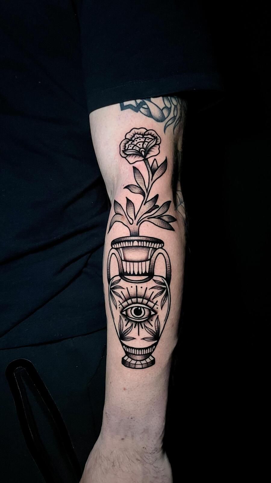 Tatuaje en blanco y negro de un jarrón con diseño de ojo y una flor en un brazo.