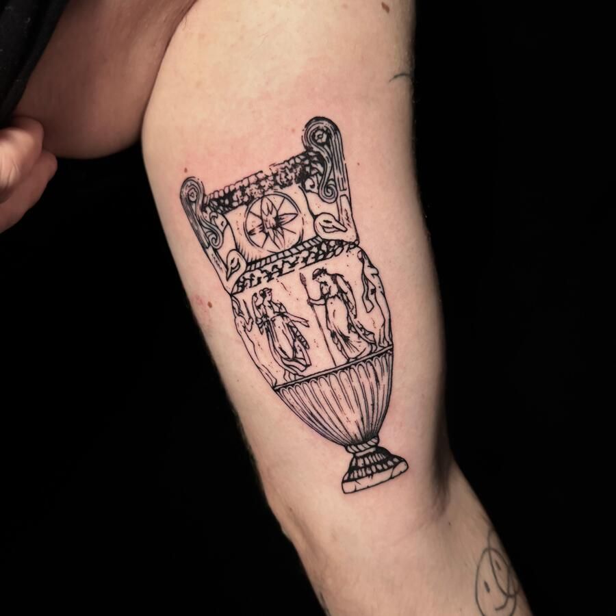 Tatuaje de tinta negra de un jarrón griego antiguo en la parte superior del brazo de una persona.