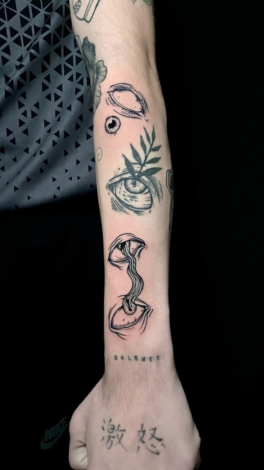 Brazo con varios tatuajes negros e indefinidos: ojos, hojas, un hongo y caracteres chinos.