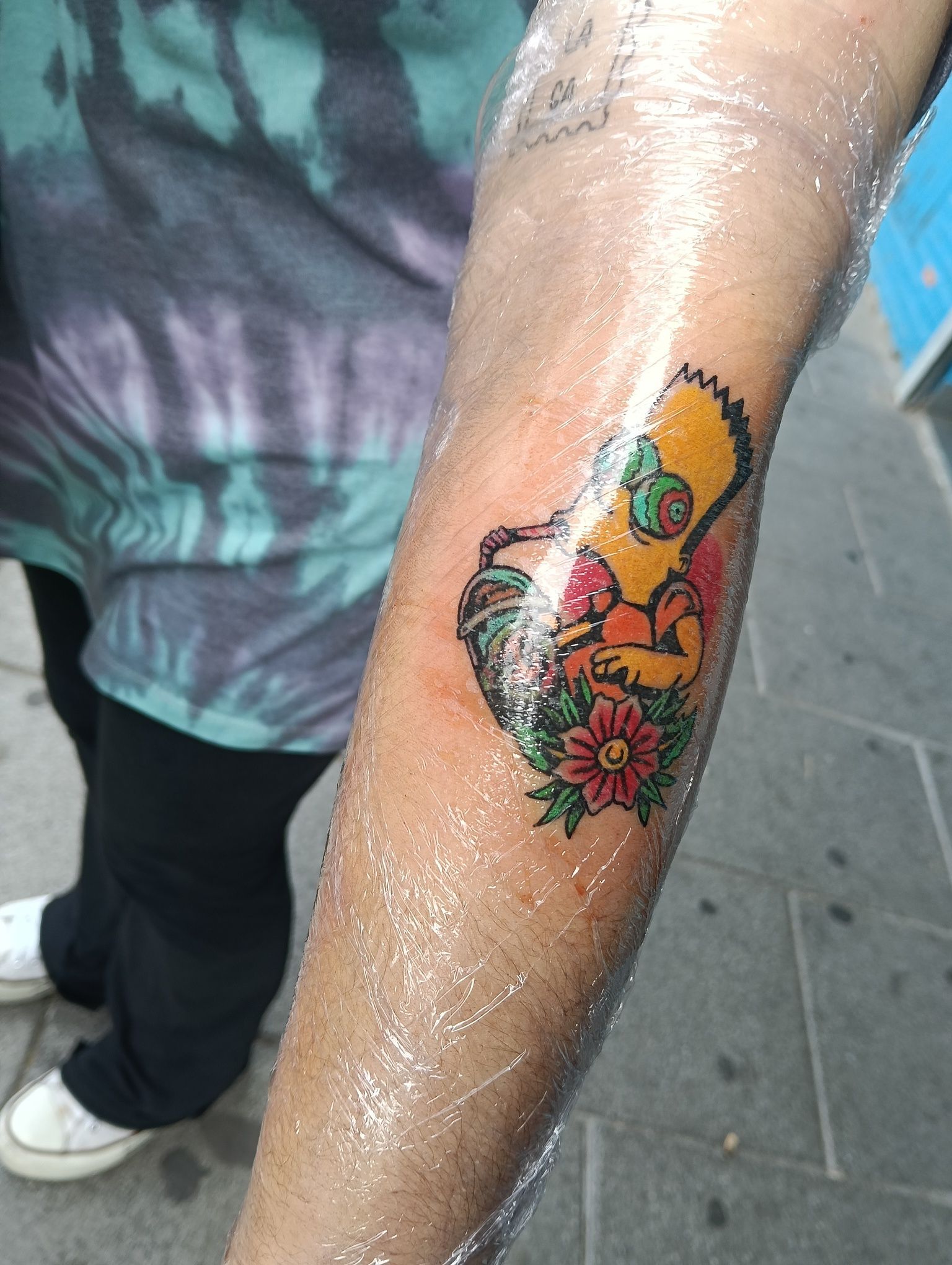 Tatuaje de Homer Simpson con flores en el brazo. El brazo tiene una sustancia brillante.