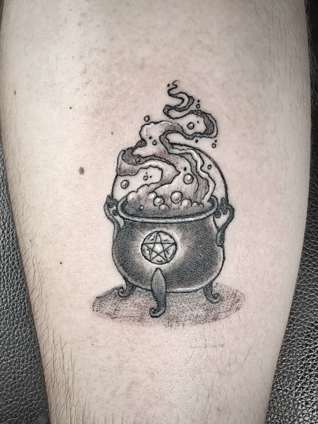Tatuaje negro y gris de un caldero burbujeante con humo y un pentagrama, sobre la piel.