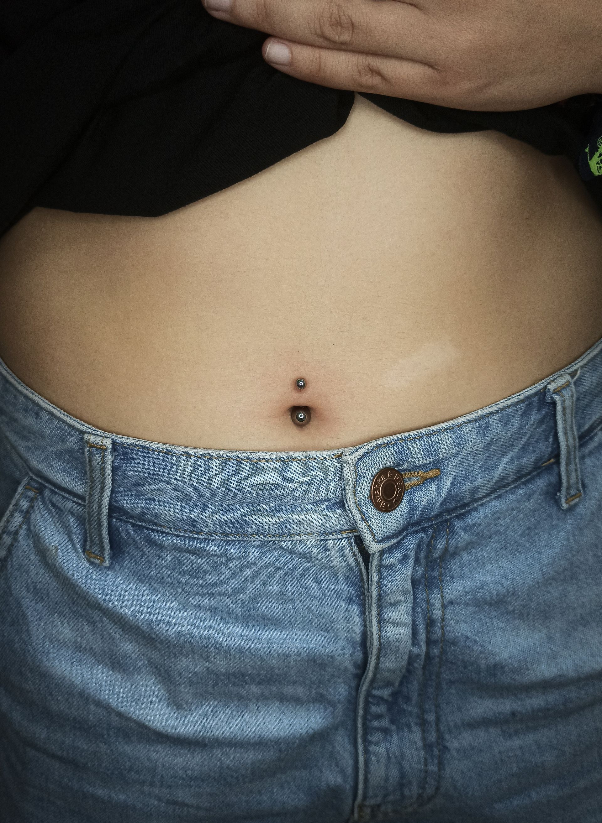 Primer plano del ombligo de una persona, perforada con una barra. Lleva camisa negra y vaqueros azules.