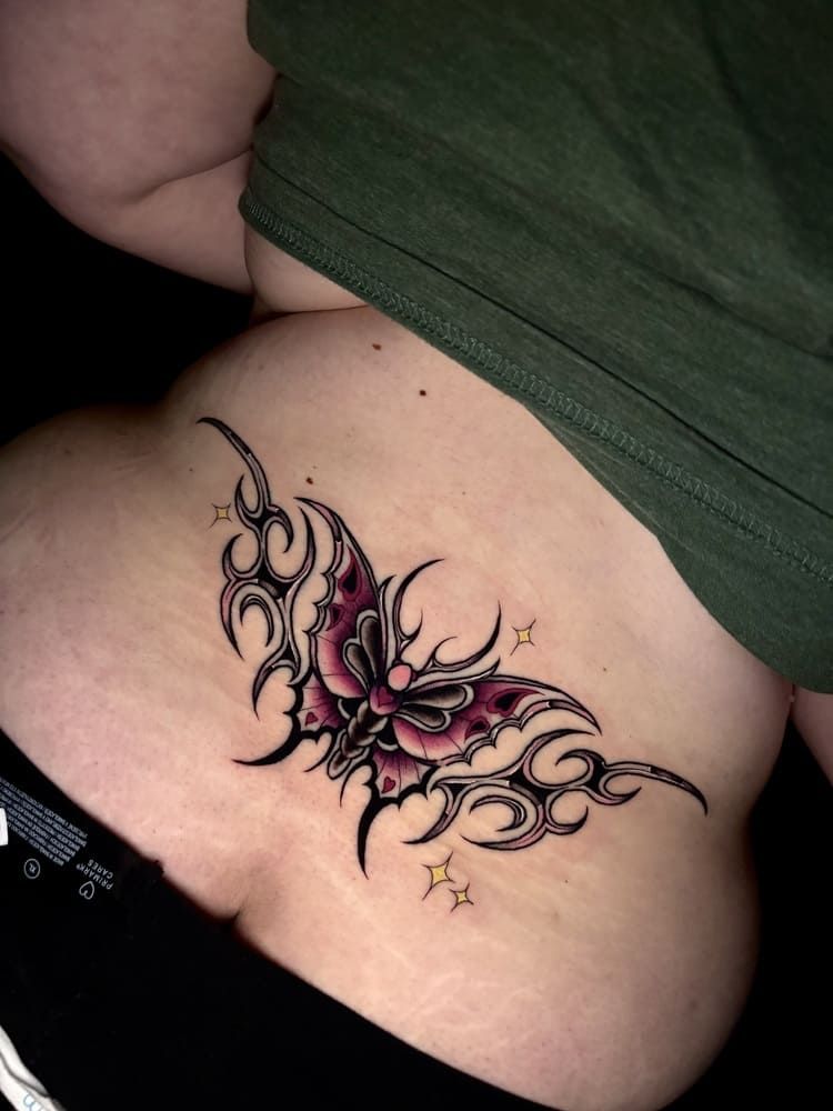Tatuaje de una mariposa negra y rosa con diseños tribales en la espalda baja de una persona, estrellas debajo.