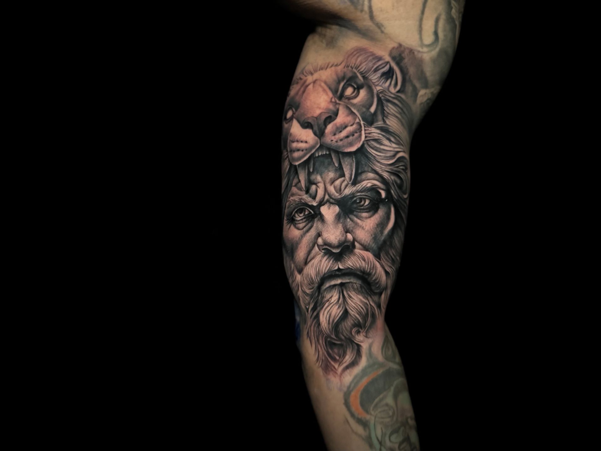Tatuaje de un hombre con expresión severa y cabeza de león en la cabeza, en un brazo oscuro.