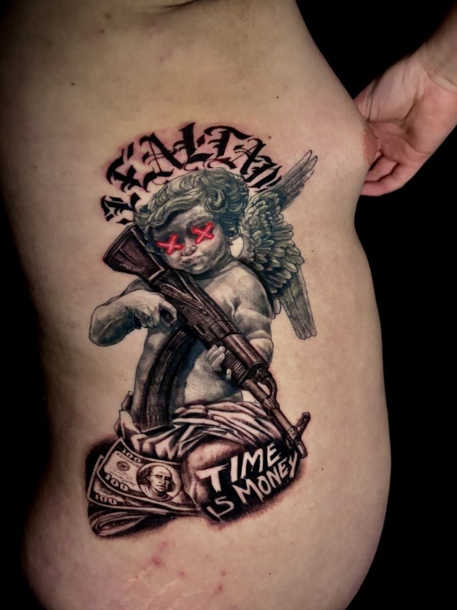 Tatuaje en el costado: ángel con un AK-47, ojos rojos y la frase 