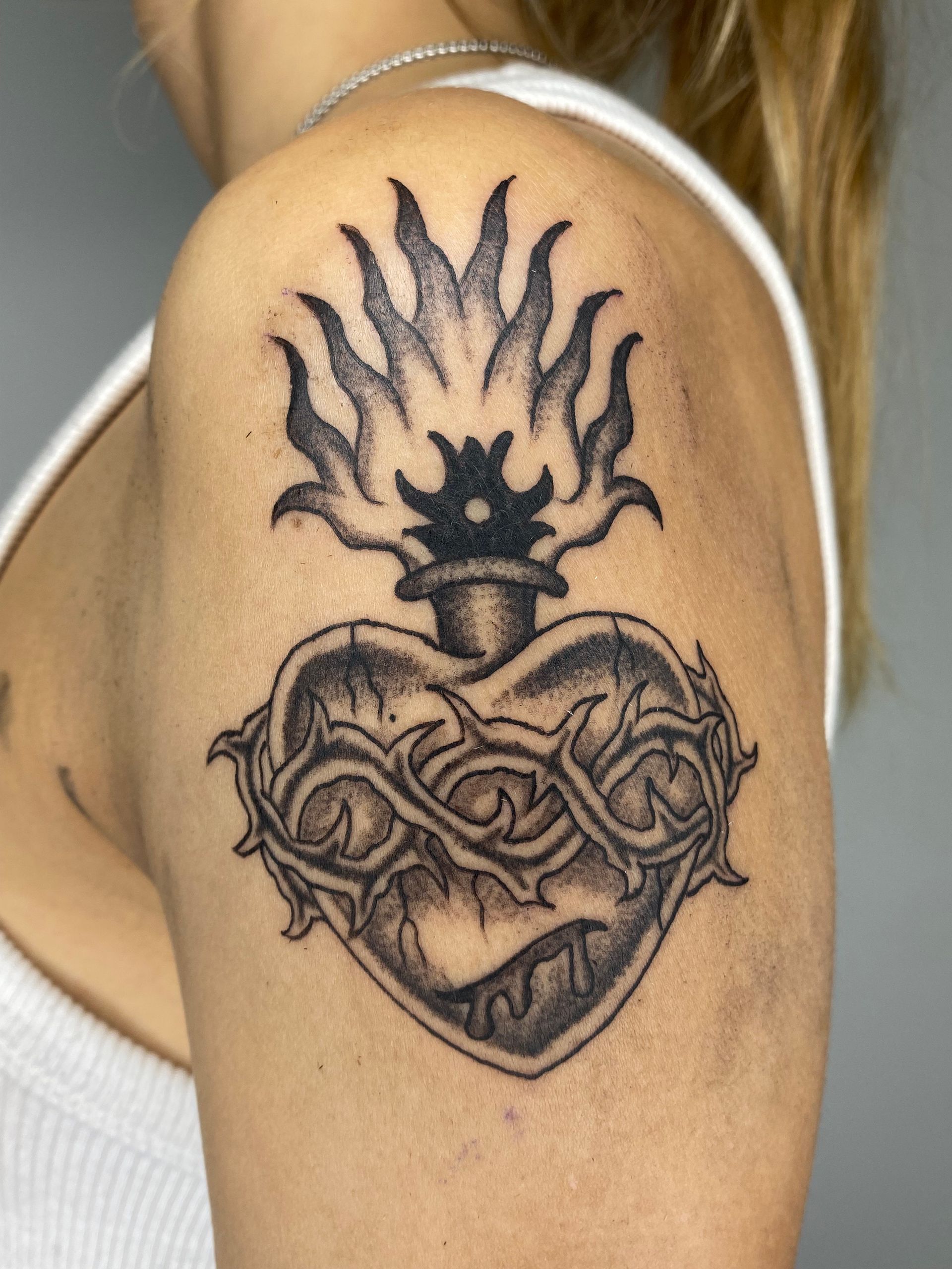 Tatuaje de un corazón con una corona de espinas y llamas en la parte superior del brazo de una persona.